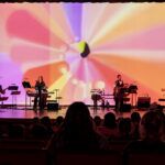 Photo of four musicians on stage with a colorful digital backdrop playing guitar, bass, keyboards with a crowd in the foreground.