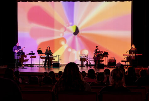 Photo of four musicians on stage with a colorful digital backdrop playing guitar, bass, keyboards with a crowd in the foreground.