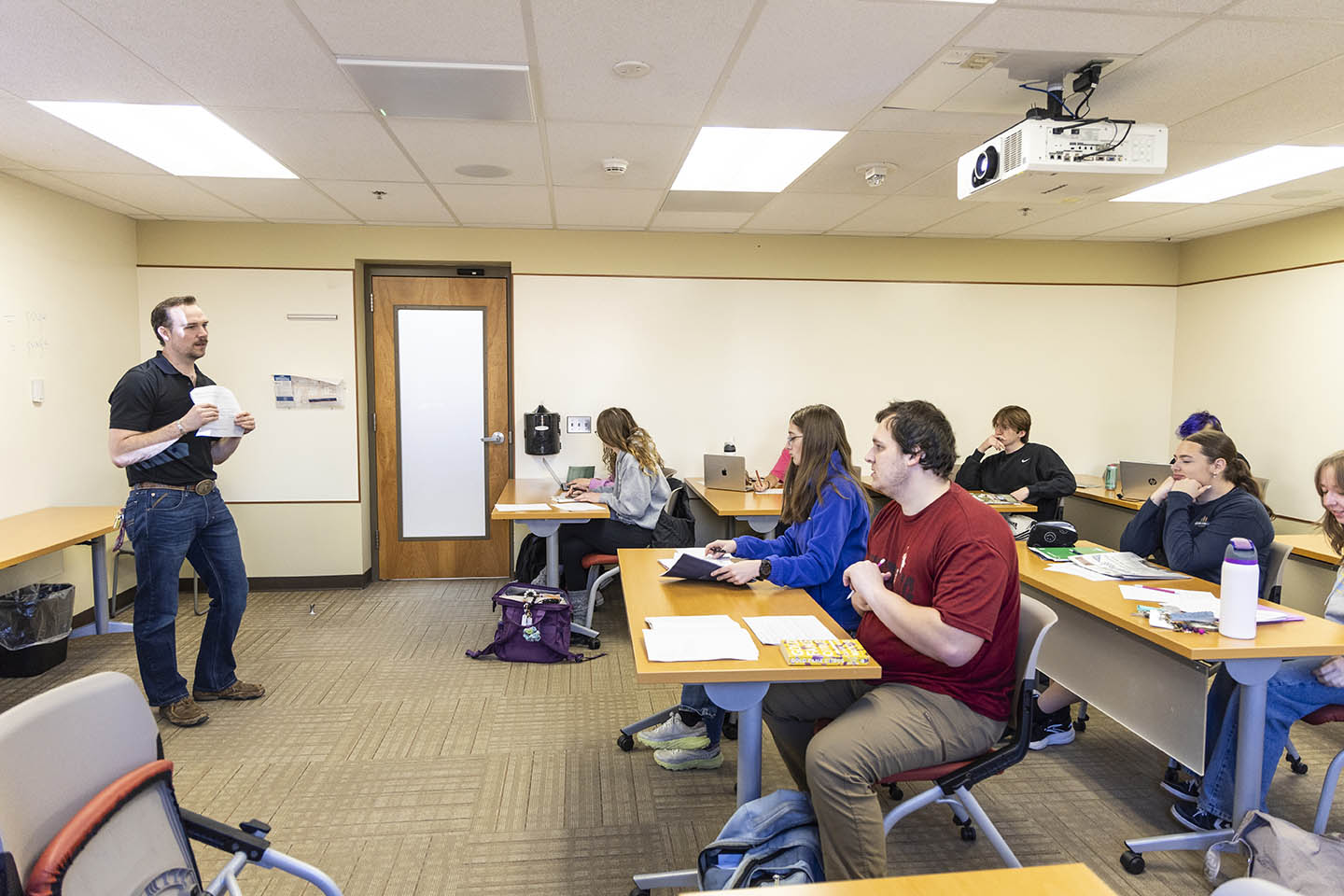 small classroom setting with students at their desks and an instructor showing them something on paper.