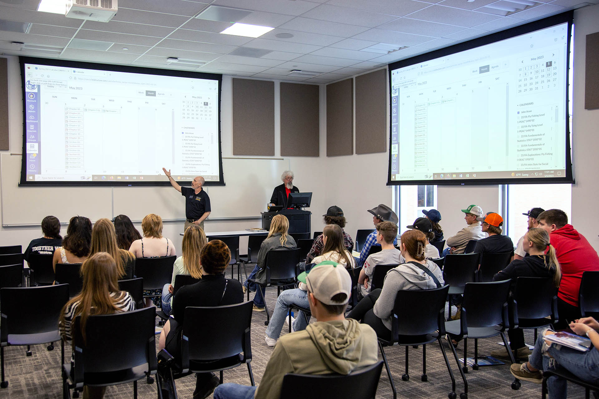 Classroom with multiple projector screens and chairs full of students watching a couple of instructors