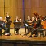 Wyoming Baroque performing on stage in Kinnison Hall with a harpsichord, upright bass, cello, viola, two violins, and a singer.