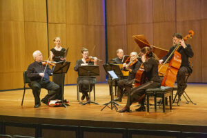 Wyoming Baroque performing on stage in Kinnison Hall with a harpsichord, upright bass, cello, viola, two violins, and a singer.