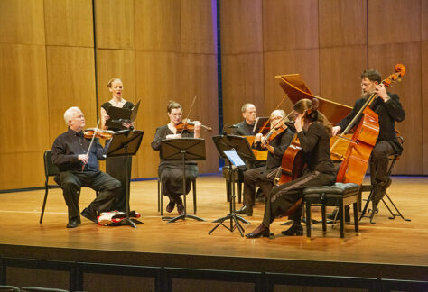 Wyoming Baroque performing on stage in Kinnison Hall with a harpsichord, upright bass, cello, viola, two violins, and a singer.