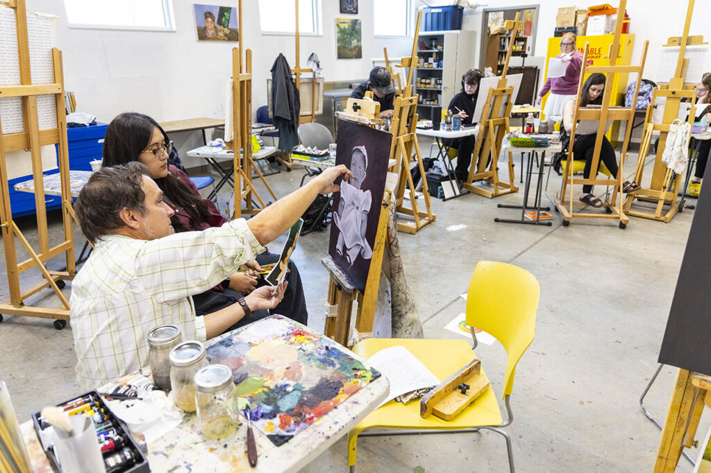 Painting and Drawing instructor David Brock works with a student on their painting on an easel in a room with other students working on other easels.