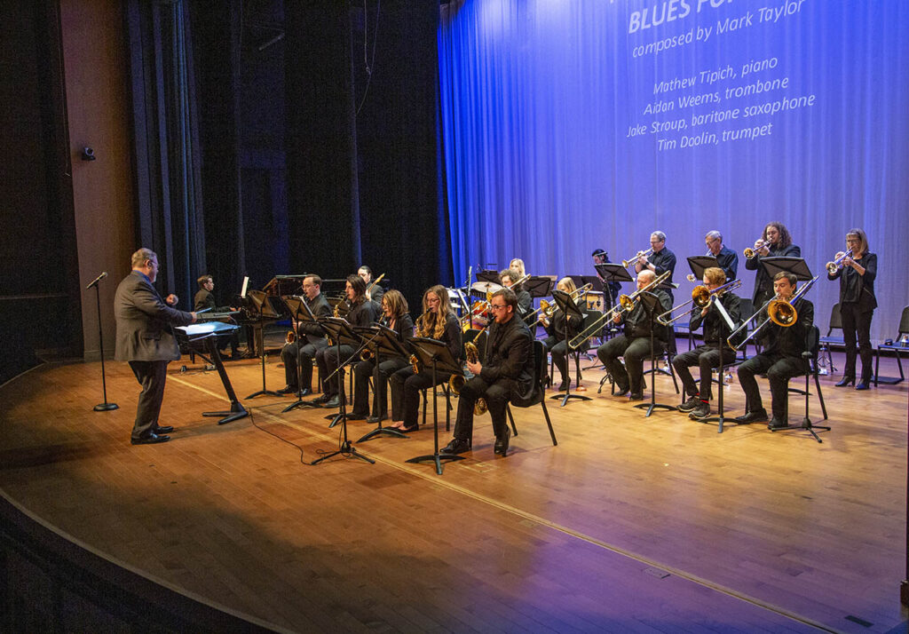 The Sheridan College Jazz Ensemble performing in Kinnison Hall, a group of 14 or so members with brass, piano, drums and electric guitar on stage.