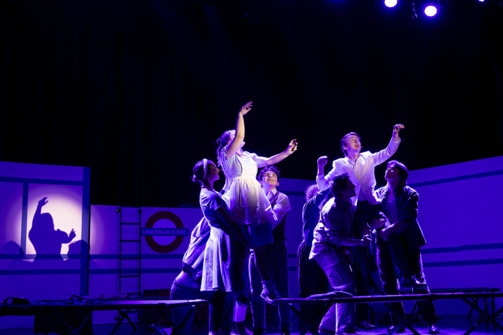 Students bathed in purple light during a theater production of alice by heart with their arms outstretched upward
