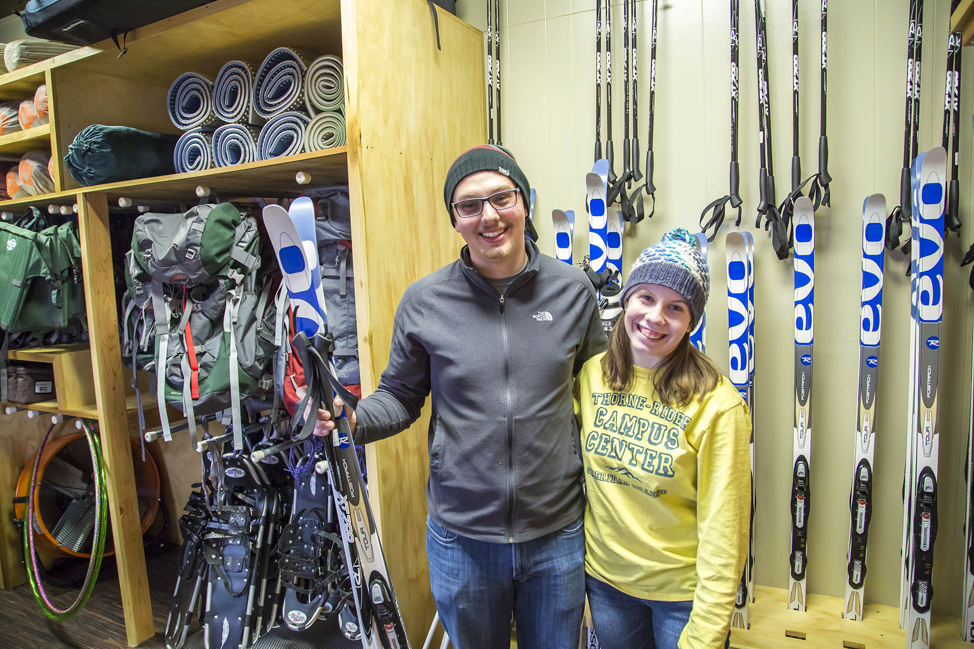 A couple students hold up their rental skis in front of the gear rack in the Innominate House