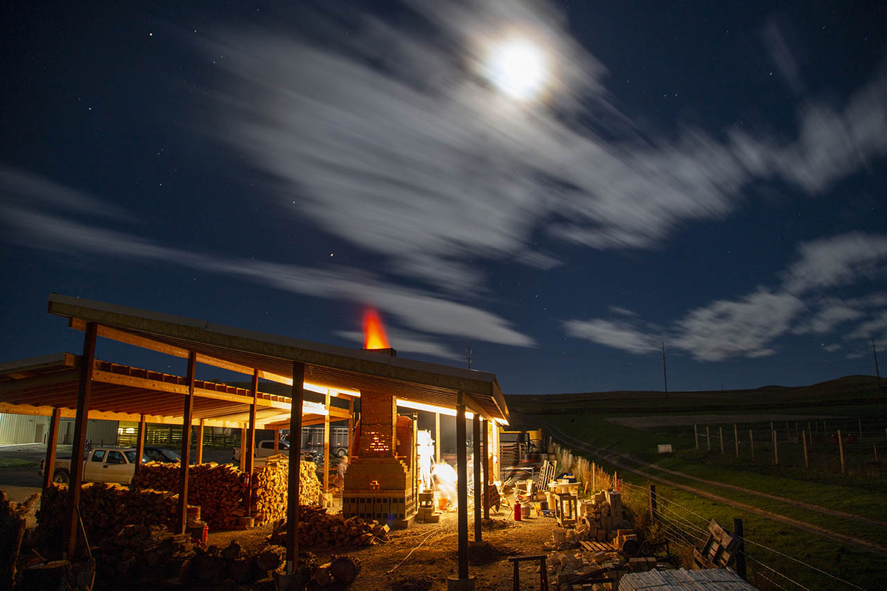 Night time image of the wood fired kiln at Sheridan College in the middle of the firing process with flames glowing out the top of the chimney into the night sky