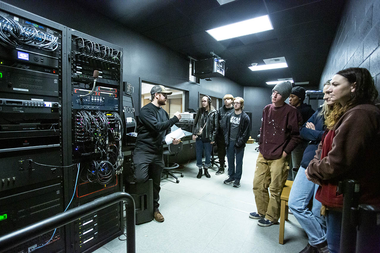 Students and technical director inside the booth in Kinnison Hall with a bunch of routing wires and patch cables visible in a tall rack tower.