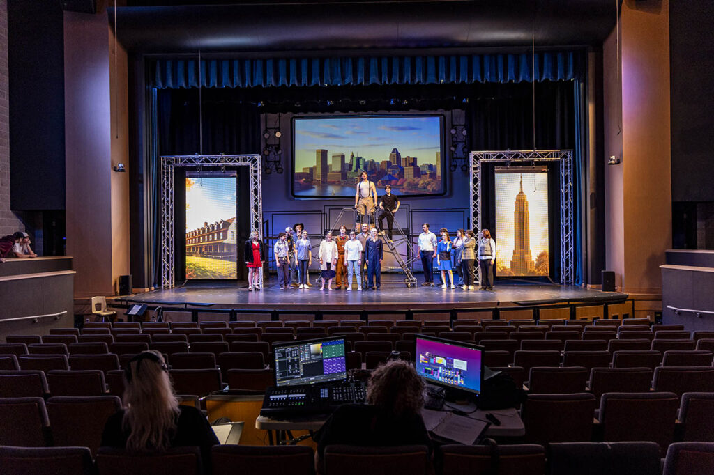 Large group of theater and dance students on stage in Kinnison Hall with colorful projection screens hanging from trusses on stage during rehearsal for the Broadway Musical Theater intensive