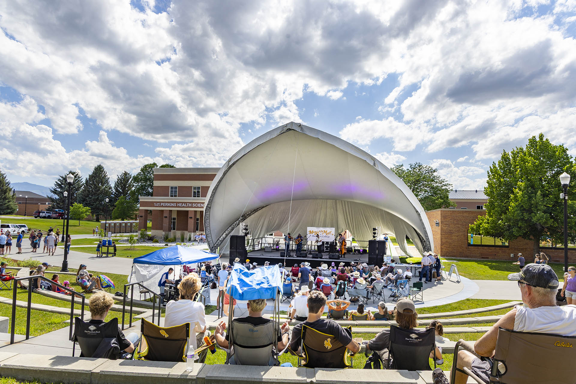 Music on the Mall showing the big tent over the stage with a string band playing and people sitting in camp chairs on the grassy terraces under a partly cloudy sky mid afternoon